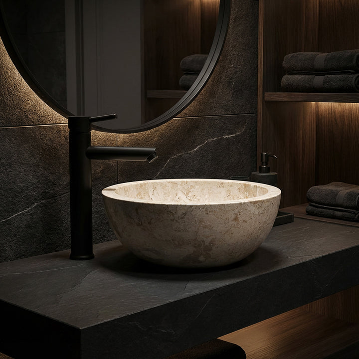 Ronde opzetwastafel van licht marmer op een donkere stenen plaat met zwart armatuur in een elegante badkamersfeer