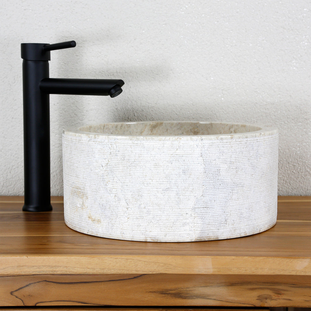 Ronde opzetwastafel van licht natuursteen met een fijne lijnstructuur, gecombineerd met een zwarte stand-alone kraan.