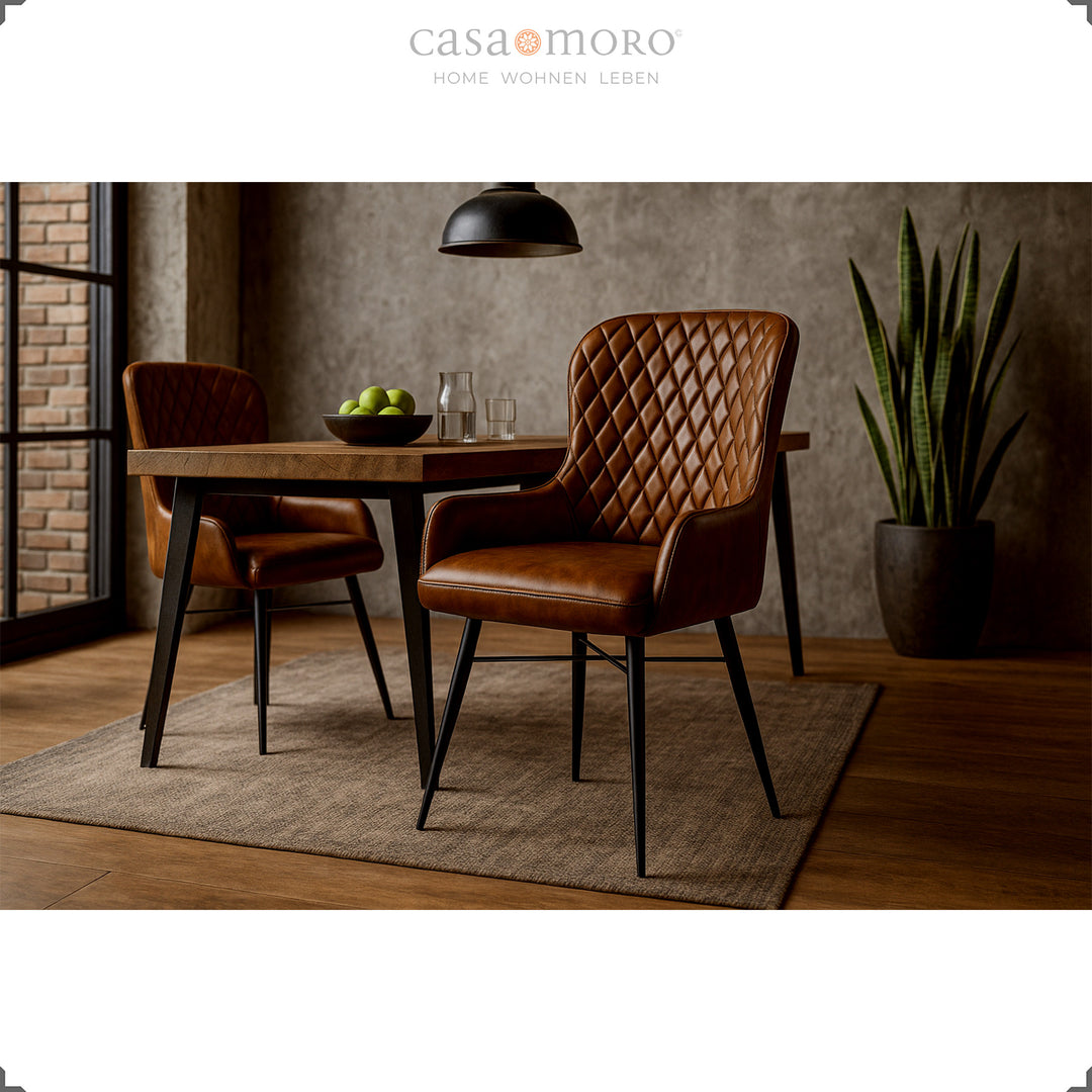 Twee antieke bruinleren stoelen met diamantquilt aan een houten tafel in de moderne eetkamer met een betonnen muur, houten vloer en groene kamerplant.