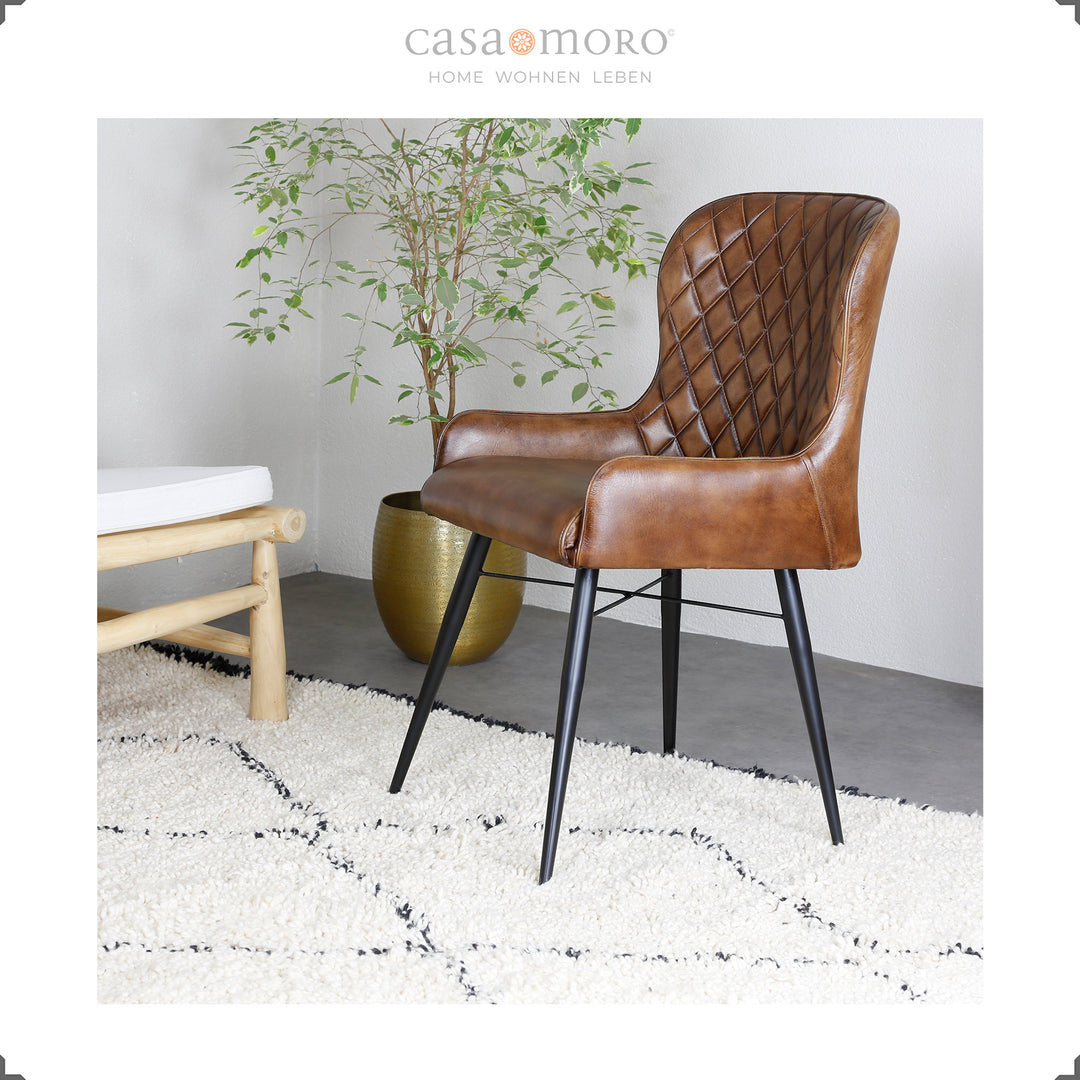 Antieke bruinleren stoel met diamantquilt op een licht tapijt, naast een gouden vaas met een groene plant - moderne woonsfeer met een natuurlijke touch.