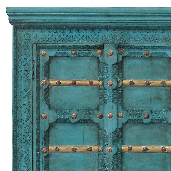 Gros plan des ornements sculptés turquoise, des traverses dorées et des ferrures sur les portes de l’armoire.