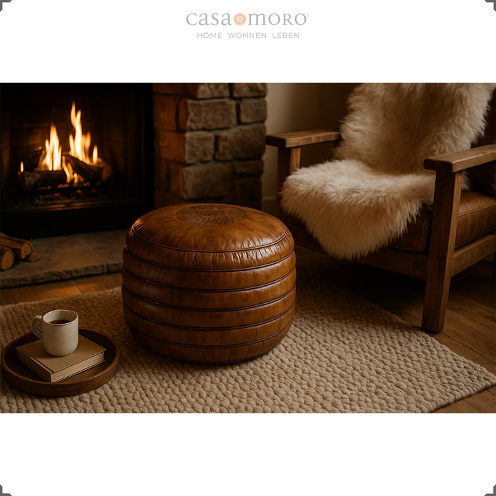 Scène de vie confortable avec un pouf rond en cuir marron chaud devant la cheminée, à côté un fauteuil en bois avec un coussin en fourrure et une tasse sur un plateau - ambiance atmosphérique.