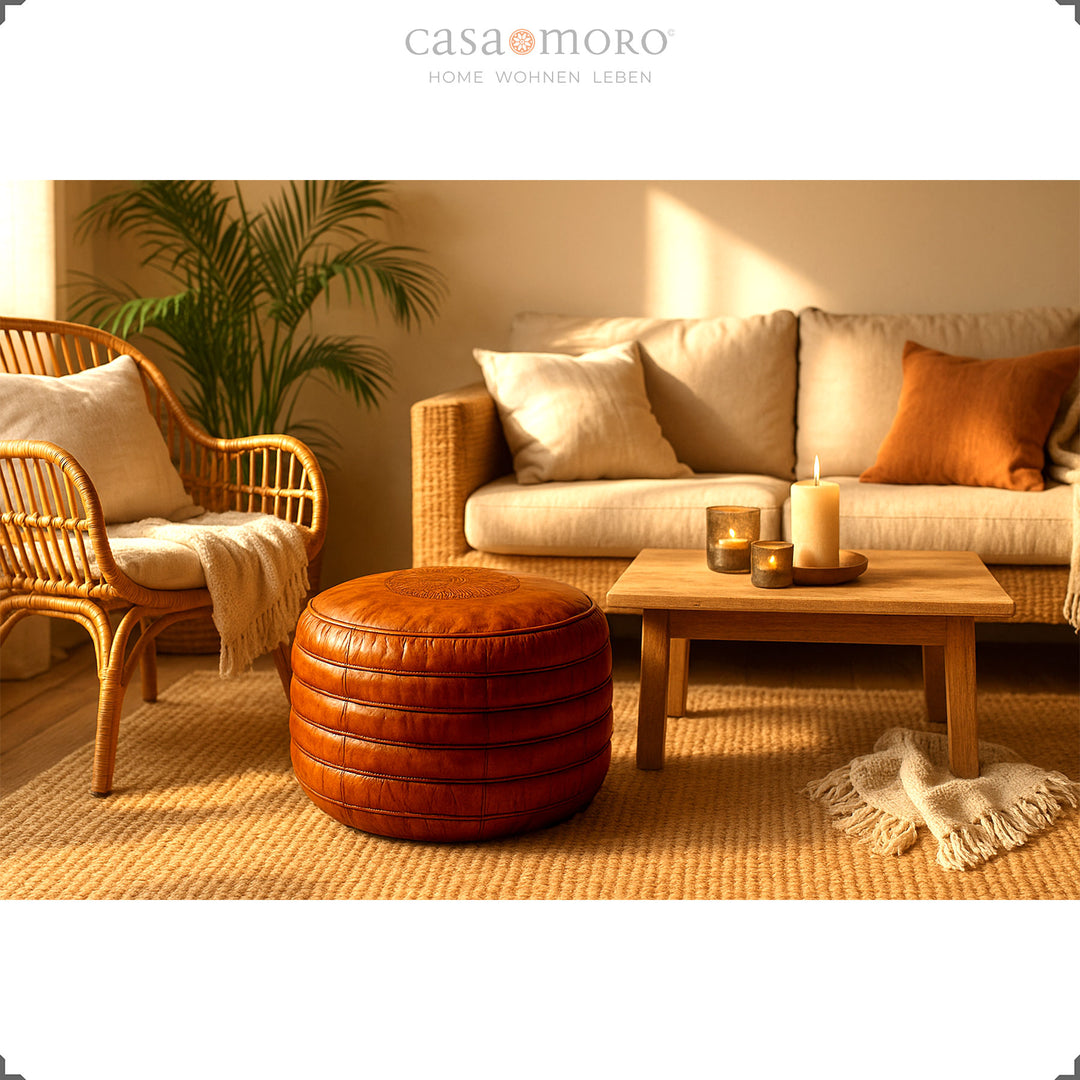Scène de vie de style bohème avec un pouf rond en cuir marron chaud devant un canapé et un fauteuil en rotin, entouré de bougies et de matériaux naturels – une ambiance harmonieuse.