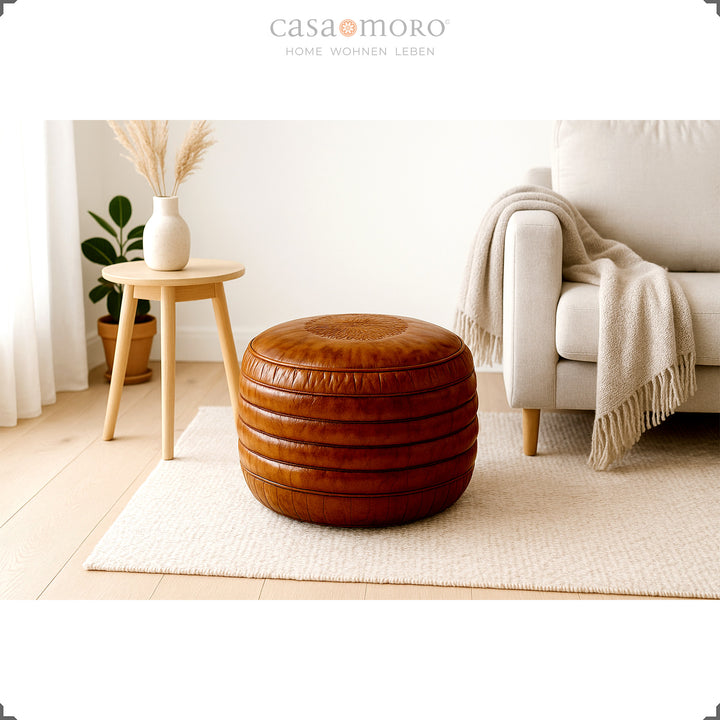 Salon lumineux de style scandinave avec un pouf rond en cuir marron sur un tapis, un canapé beige et un tabouret en bois avec un vase : une ambiance calme et naturelle.