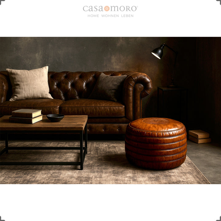 Salon élégant avec canapé Chesterfield, table basse et pouf rond en cuir marron - scène élégante dans un style industriel et moderne-rustique.