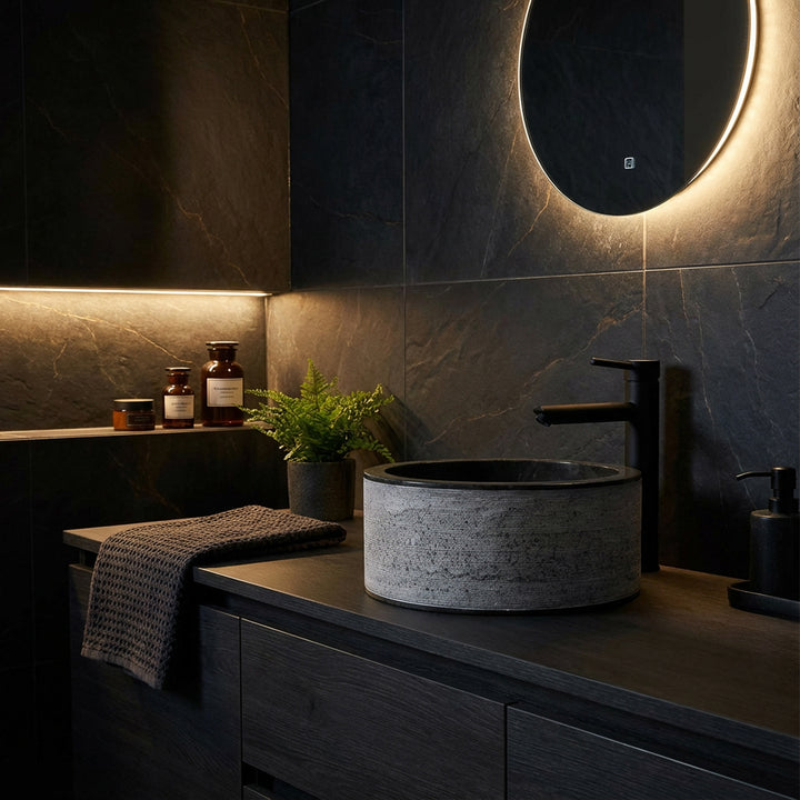 Ronde stenen wastafel met fijne structuur op een donkerhouten console, sfeervol geënsceneerd in de moderne dark mood badkamer.