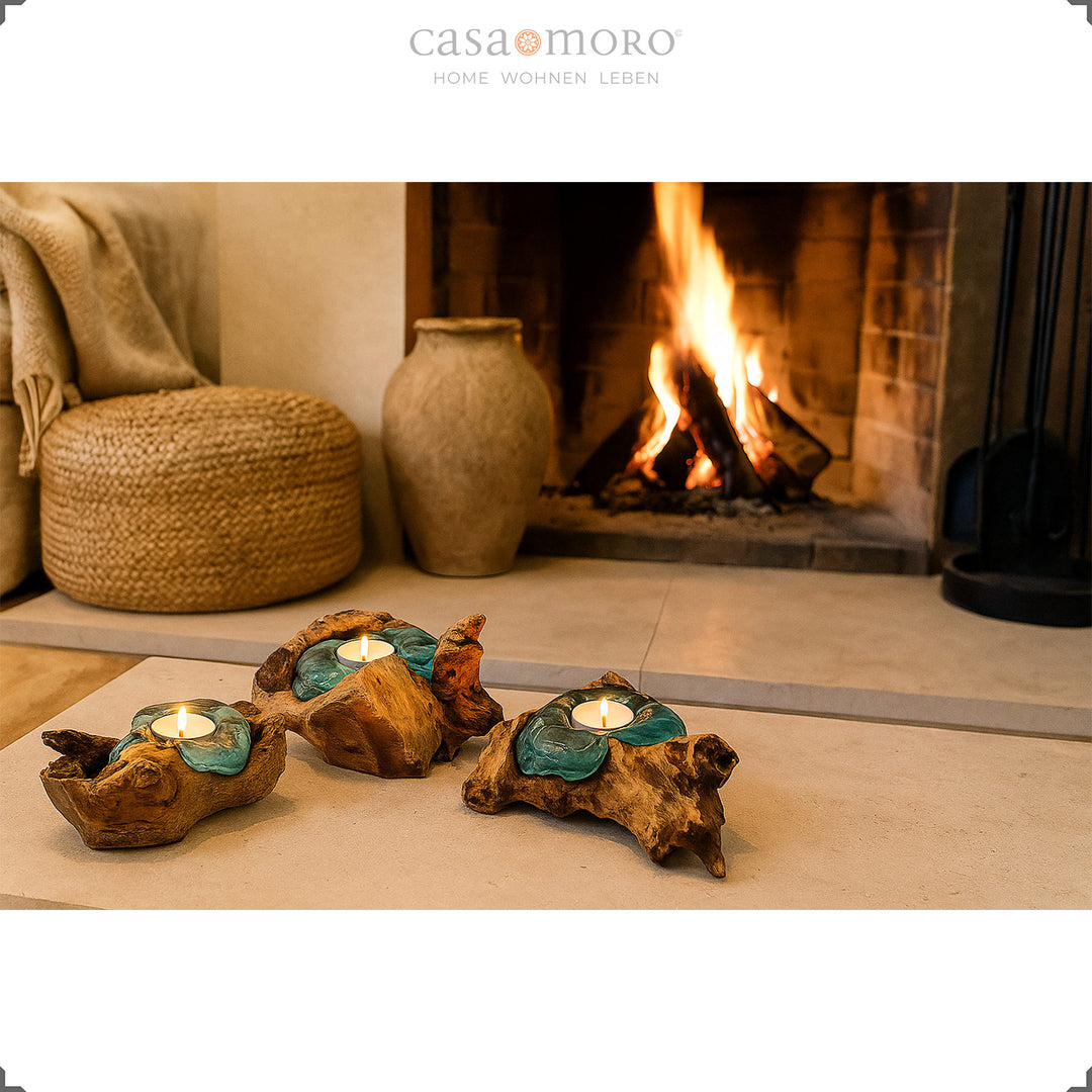 Drie theelichthouders van glas en wortelhout voor een brandende open haard, hun zachte licht onderstreept de gezellige woonsfeer.
