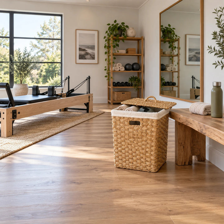 Panier carré en jonc de mer, serviettes et couvercle posé en biais, près d’un banc en bois dans la salle de Pilates.