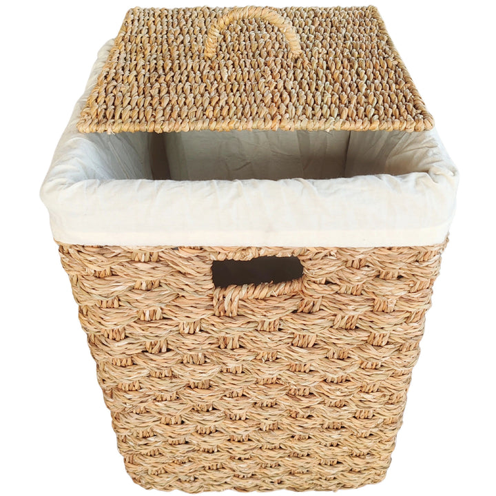 Panier à linge carré en jonc de mer, couvercle soulevé en biais, sac clair et ouverture de prise à l’avant.