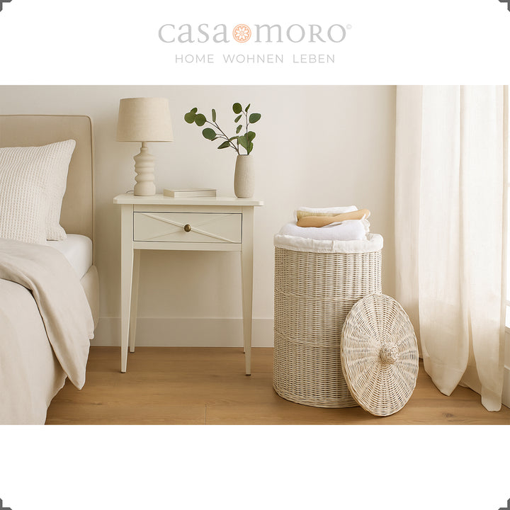 Panier à linge blanc brillant dans la chambre, combiné avec des tons naturels chauds, des rideaux et une table de chevet