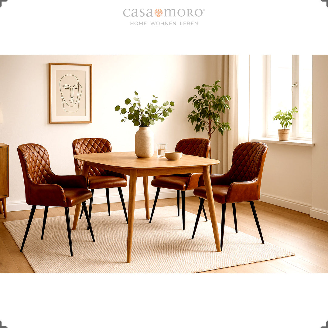 Vier antieke bruinleren stoelen met diamantquilt rond een ronde houten tafel in een lichte, vriendelijke woonkamer met planten en natuurlijk licht.